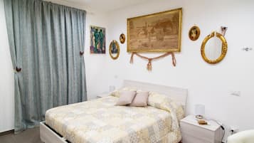 Una camera da letto con un letto, un comodino, una sedia, una finestra con tende e decorazioni a parete tra cui quadri e specchi.