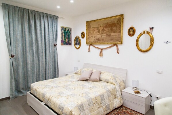 A bedroom with a bed, a nightstand, a chair, a window with curtains, and wall decorations including paintings and mirrors. - First House Posillipo Holiday Home Naples (Naples)