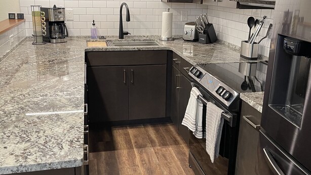 A modern kitchen with dark cabinets, a stainless steel refrigerator, a built-in oven, and a granite countertop.
