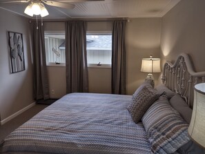 Une chambre à coucher avec un lit, un ventilateur de plafond, une fenêtre avec des rideaux, une lampe fixée au mur et un tableau encadré.