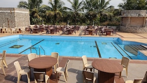 A poolside area with multiple seating arrangements, including tables and chairs, surrounded by a stone wall and palm trees. - Kanthi Resorts Badami (Bagalkot)