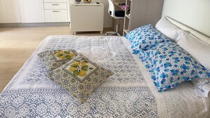 A bedroom with a bed, pillows, a bedside table, a chair, and a small desk with a computer. There is a kitchen area in the background with cabinets and a microwave. - Room-the Archi B&B Apartment (Anacapri)