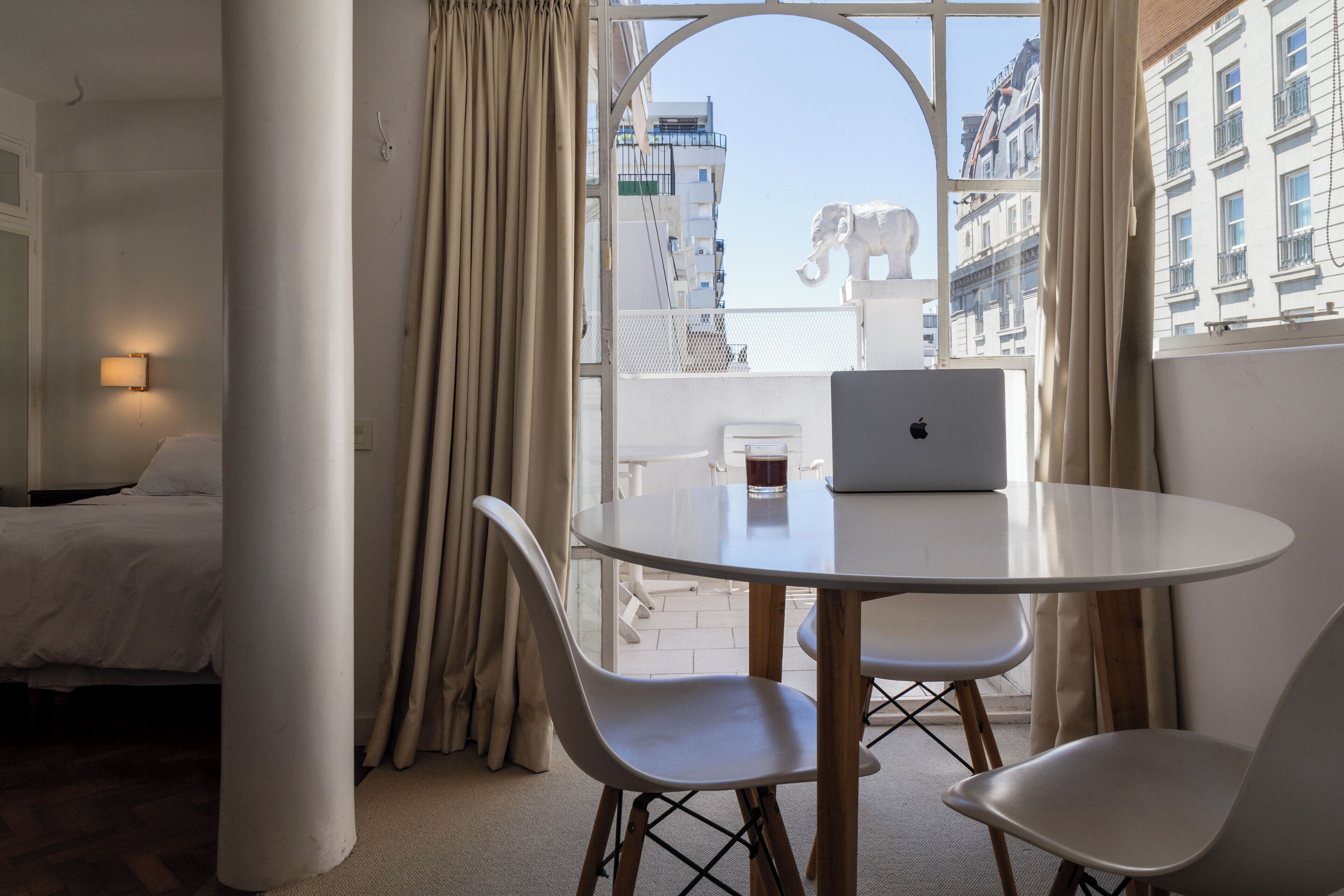 Une chambre d’hôtel avec un lit, une table ronde sur laquelle se trouve un ordinateur portable, et une vue sur les bâtiments à travers de grandes fenêtres.