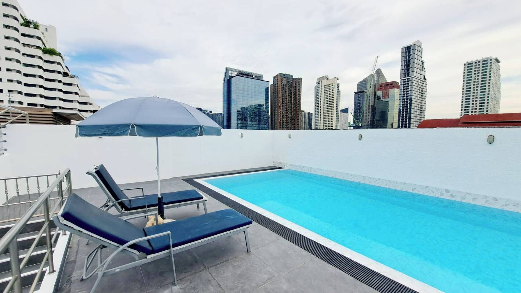 A rooftop swimming pool with lounge chairs and an umbrella, surrounded by high-rise buildings. - 5A - 2 Bedroom Downtown Apartment With Shared Pool (Bangkok)