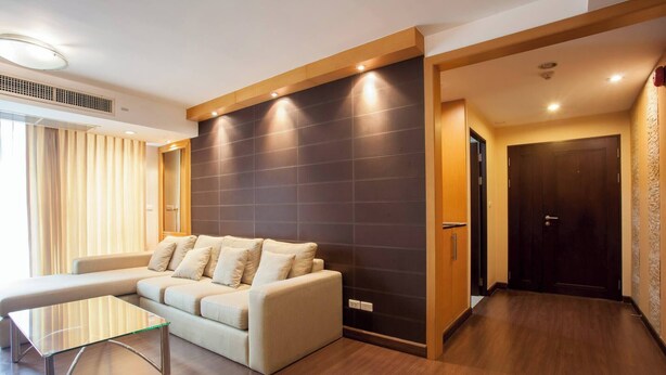 A modern living room with a sectional sofa, a glass coffee table, and a dark tiled accent wall. - 5A - 2 Bedroom Downtown Apartment With Shared Pool (Bangkok)