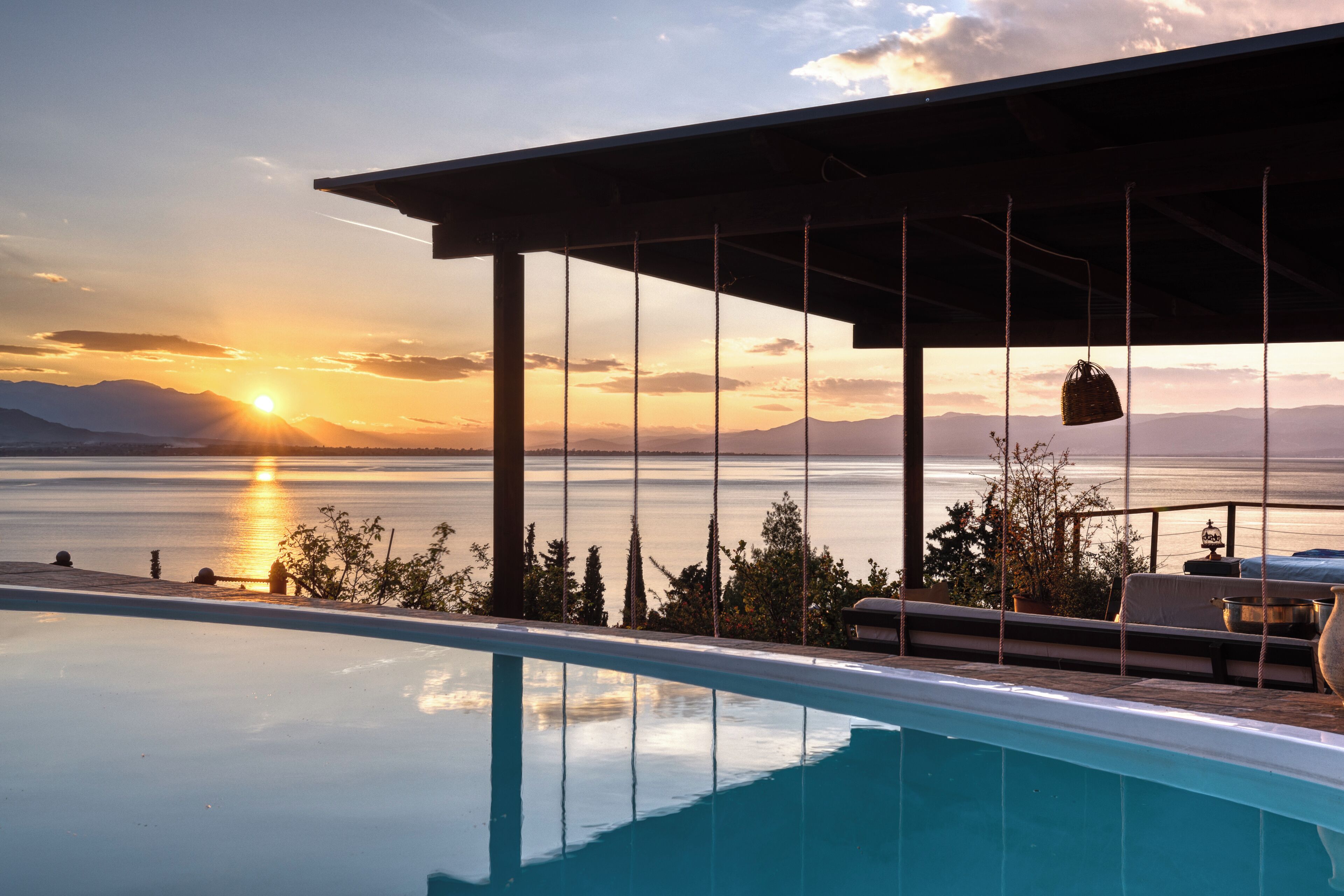 An infinity pool with a view of the sunset over a body of water, a pergola structure, and outdoor furniture.