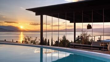 An infinity pool with a view of the sunset over a body of water, a pergola structure, and outdoor furniture.