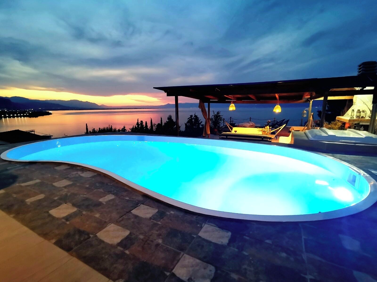 Una piscina con borde curvo, rodeada de baldosas de piedra. Hay una zona cubierta con asientos y luces colgantes, y se observa un lago y montañas al fondo.