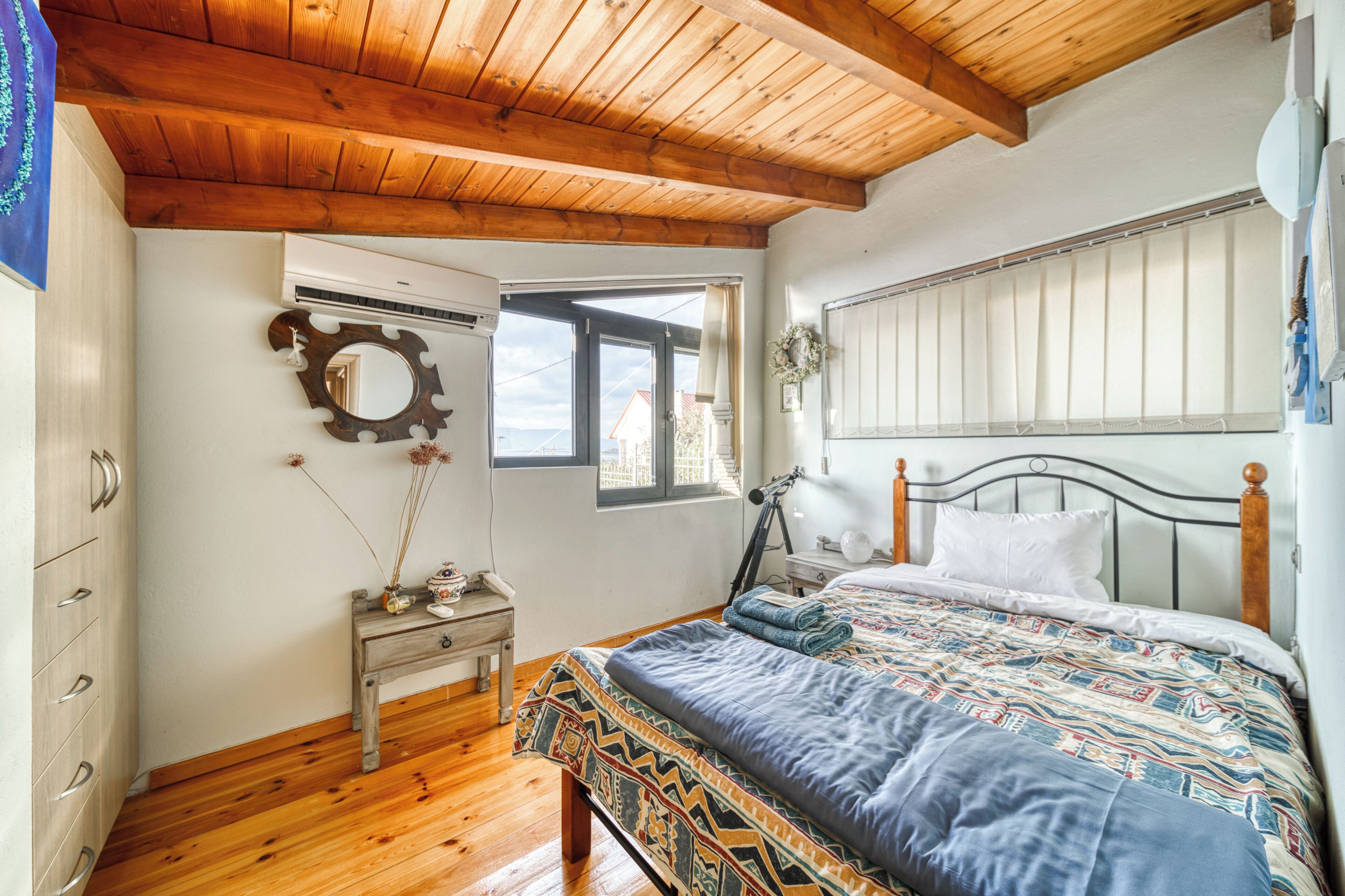A bedroom with a wooden ceiling, a bed with a patterned bedspread, a nightstand with a lamp, and a window with a view of the outside.