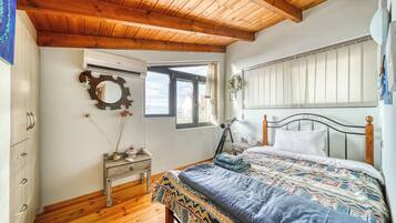 A bedroom with a wooden ceiling, a bed with a patterned bedspread, a nightstand with a lamp, and a window with a view of the outside.