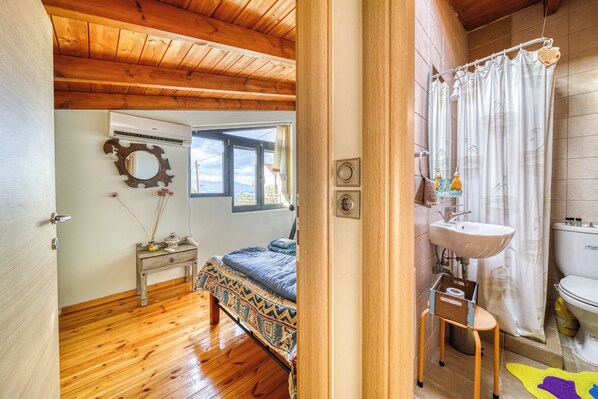 A room with wooden flooring, a bed with a patterned bedspread, a small bedside table with a mirror, a window with a view, and a bathroom with a sink and toilet.