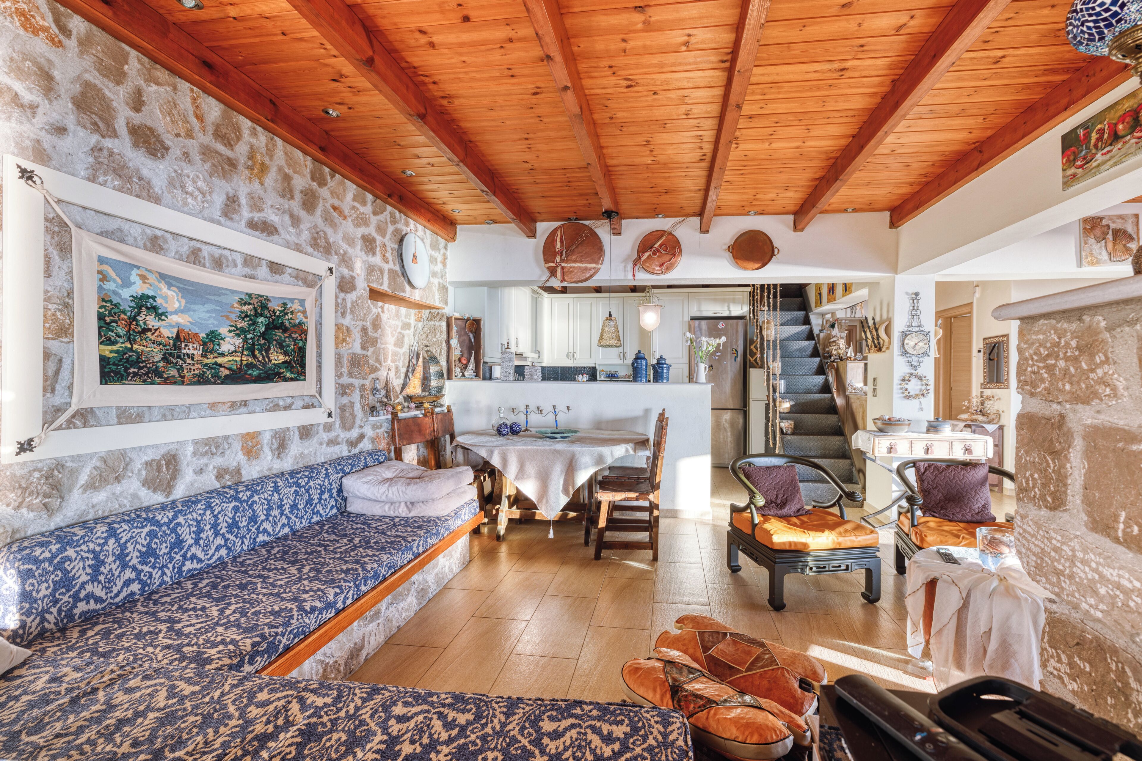 A cozy interior with a stone wall, wooden ceiling beams, a dining area with a table and chairs, and a staircase leading to an upper level.