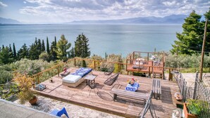 Eine Holzterrasse mit Gartenmöbeln, einem Tisch mit blauer Tischdecke und Blick auf einen See und Berge.