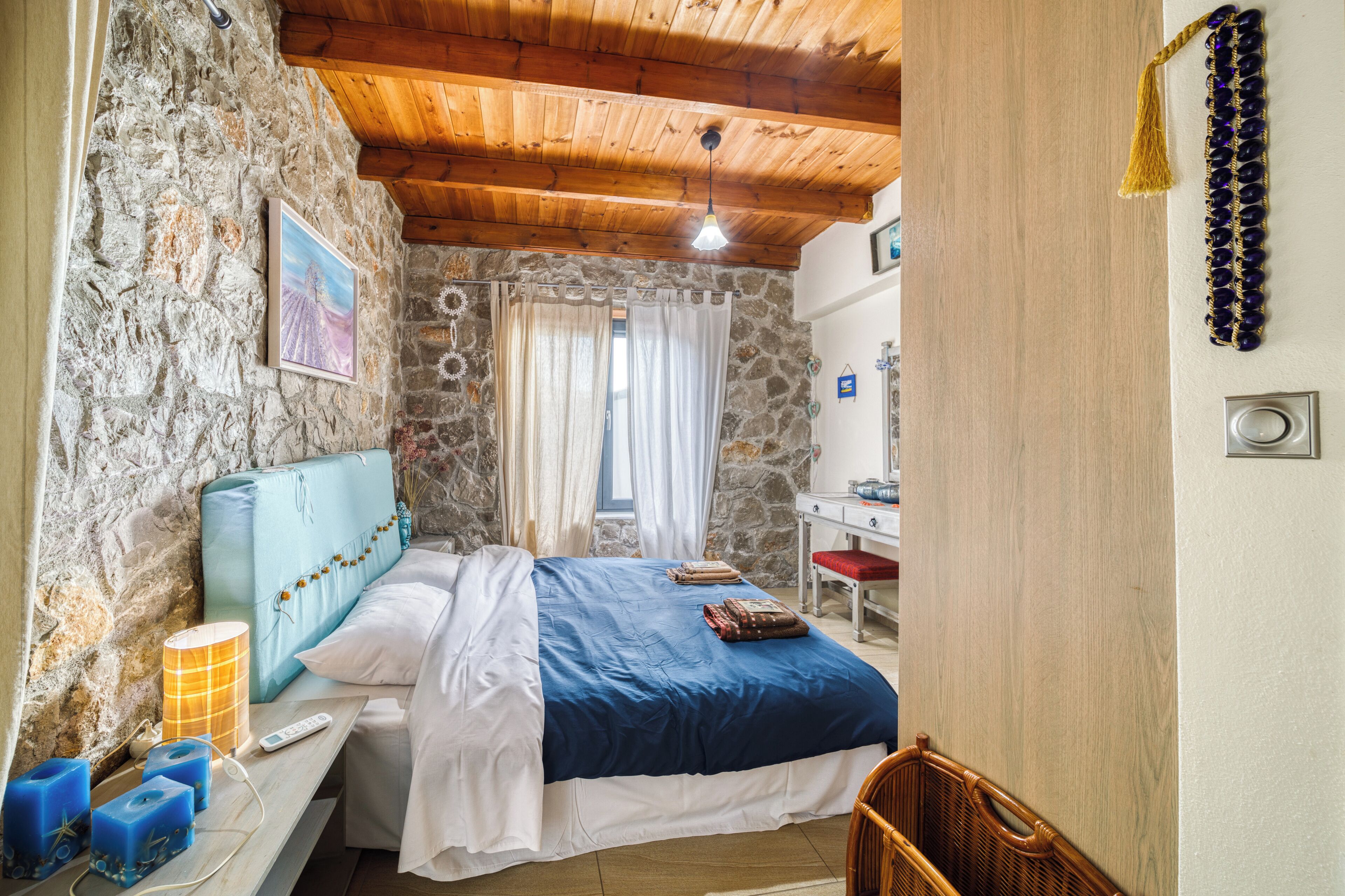 Une chambre avec un mur en pierre, un plafond en bois, un lit recouvert d’une couvre-lit bleue et blanche, une table de chevet avec une lampe, et un petit bureau avec une chaise.