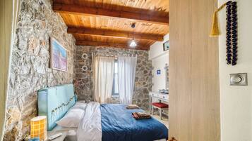 Une chambre avec un mur en pierre, un plafond en bois, un lit recouvert d’une couvre-lit bleue et blanche, une table de chevet avec une lampe, et un petit bureau avec une chaise.