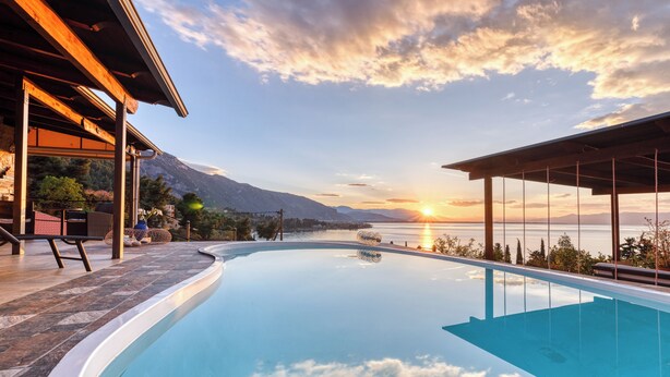 Ein Swimmingpool mit Blick auf einen Sonnenuntergang über dem Wasser, eine überdachte Terrasse und eine Berglandschaft im Hintergrund.
