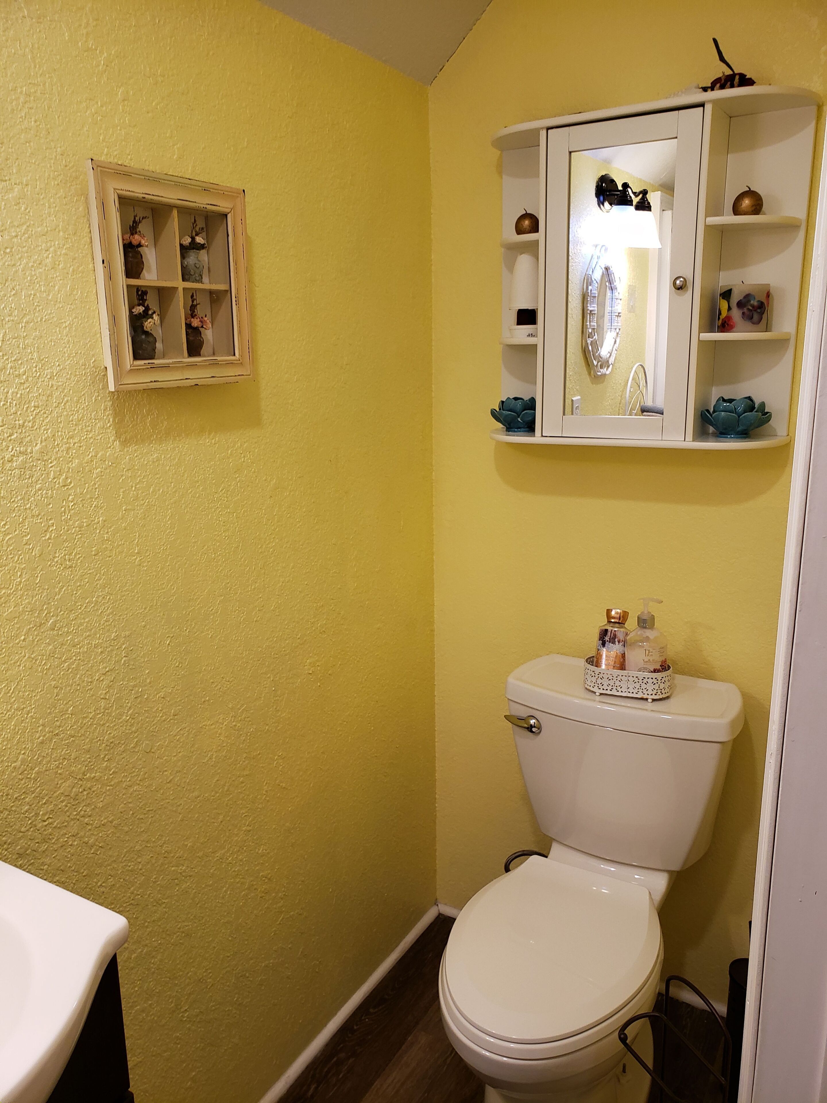 A bathroom with a yellow wall, a white toilet, a small cabinet with a mirror, and a shelf with toiletries.