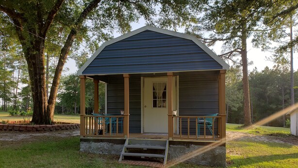 A small, single-story cabin with a porch and a door, surrounded by trees and grass.