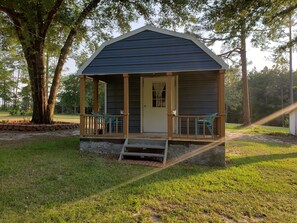 Een klein, gelijkvloers blokhut met een veranda en een deur, omringd door bomen en gras.