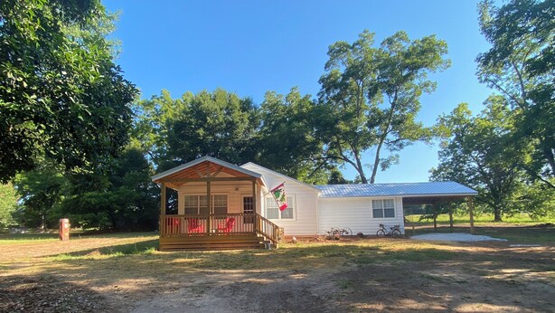 A small cabin with a porch and a covered area, surrounded by trees and a dirt path.