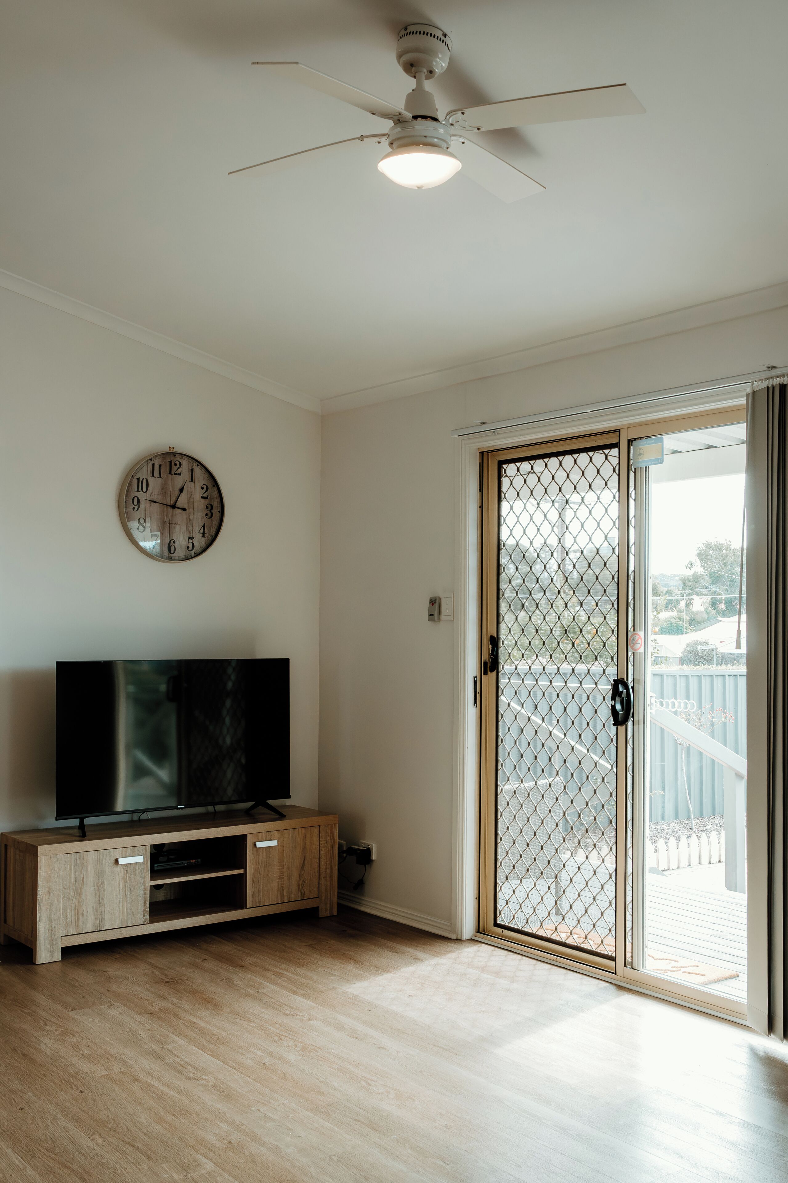 A room with a ceiling fan, a TV on a wooden cabinet, a sliding glass door, and a wall clock.