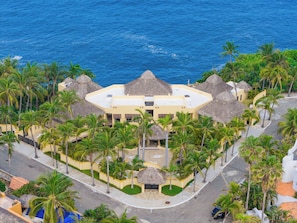 A resort with a central building surrounded by palm trees and thatched-roof structures, overlooking a body of water. - The Ultimate Indulgence (Huatulco)