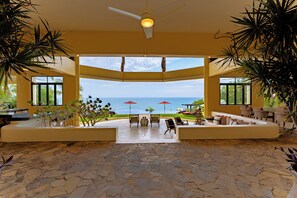 A spacious indoor area with a view of the ocean, featuring a ceiling fan, outdoor seating, and potted plants. - The Ultimate Indulgence (Huatulco)