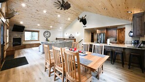 Een ruime keuken met houten kasten, een eethoek met houten stoelen, een open haard en een grote klok aan de muur.