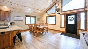 Een chaletinterieur met een houten eettafel, stoelen, een keukenhoek en een groot raam.