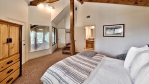 A bedroom with a large bed, wooden ceiling beams, a view through a window, and a door leading to another room.