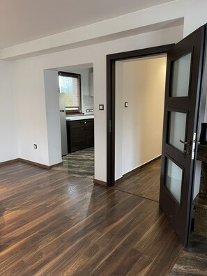 A modern interior with a wooden floor, a kitchen area with a window, and a door leading to another room.
