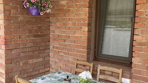 Un petit espace patio avec un mur en briques, une fenêtre, une table recouverte d’une nappe à motifs de feuilles, deux chaises en bois, un pot de fleurs suspendu et un petit récipient contenant une cuillère.