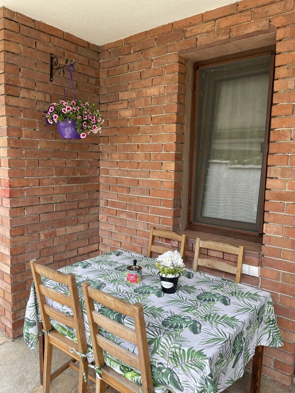 A small patio area with a brick wall, a window, a table with a leaf-patterned tablecloth, two wooden chairs, a hanging flower pot, and a small container with a spoon.