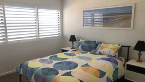 Un dormitorio con una cama, dos mesitas de noche, una lámpara en cada una y un cuadro de una escena de playa en la pared.