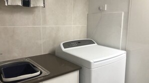 A compact, white washing machine with a control panel, situated next to a small sink with a dark basin. - Kings Beach Apt: Ocean Views & Rooftop (KINGS BEACH)