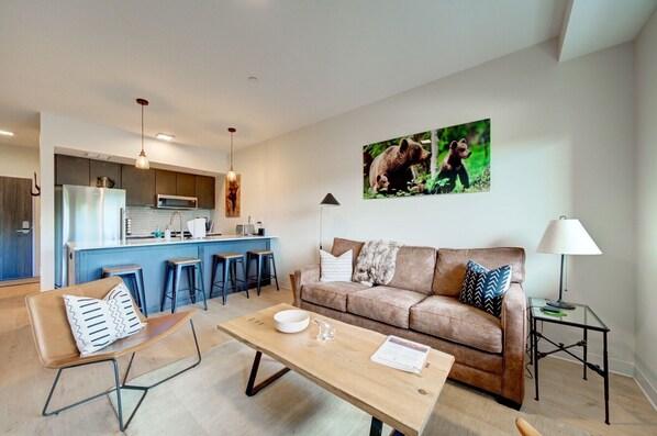 A modern living room with a sofa, coffee table, and bar stools. A kitchen with a refrigerator and microwave is visible in the background.