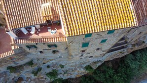 Vue aérienne d’un bâtiment historique en pierre, doté d’un toit en tuiles, d’une cour et d’un mur de pierre abrupt surplombant une zone boisée.