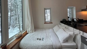 A bedroom with a large window, a bed with white bedding, a bedside table, and a small window. - Black Spruce - Red Door  (Whitehorse)