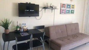 Ein Wohnzimmer mit Flachbildfernseher, einer Couch, einem Esstisch mit Stühlen, einem kleinen Regal für elektronische Geräte und einer Topfpflanze.