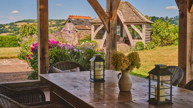 Uma pérgula de madeira com uma mesa e cadeiras, rodeada por flores e uma vista para casas ao longe.