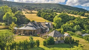 Uma casa grande com um alpendre amplo e um gazebo, cercada por vegetação exuberante e colinas ondulantes.