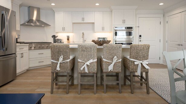 A modern kitchen with white cabinetry, stainless steel appliances, and a central island with four wicker chairs tied with white ribbons.