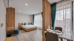 Una habitación de hotel moderna con una cama de cabecera de madera, una mesa de comedor con sillas y ventanas grandes con cortinas.