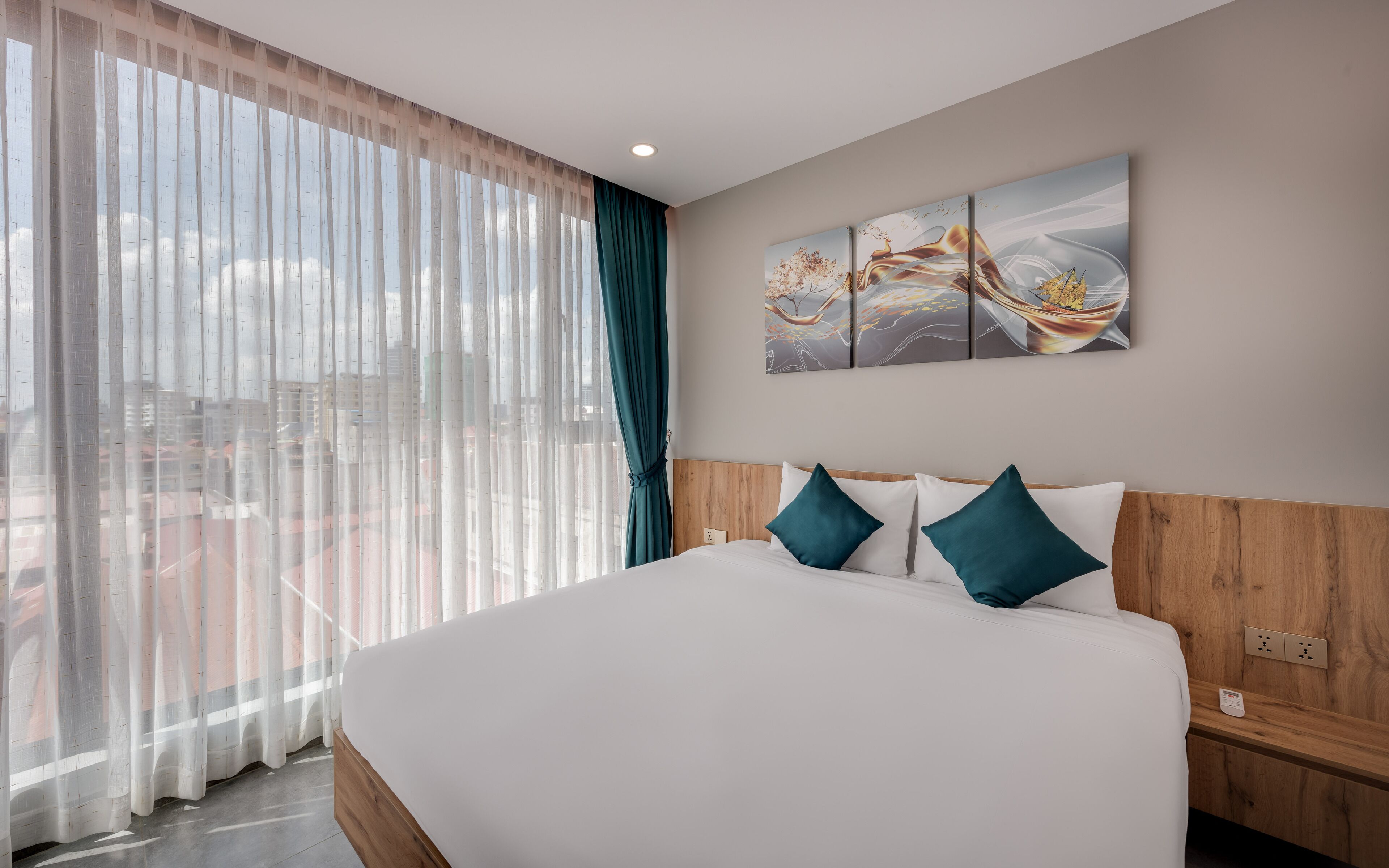A hotel room with a large bed, a wooden headboard, a view of the city through sheer curtains, and a wall-mounted abstract art piece.
