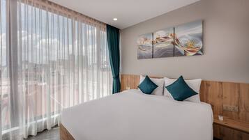 A hotel room with a large bed, a wooden headboard, a view of the city through sheer curtains, and a wall-mounted abstract art piece.