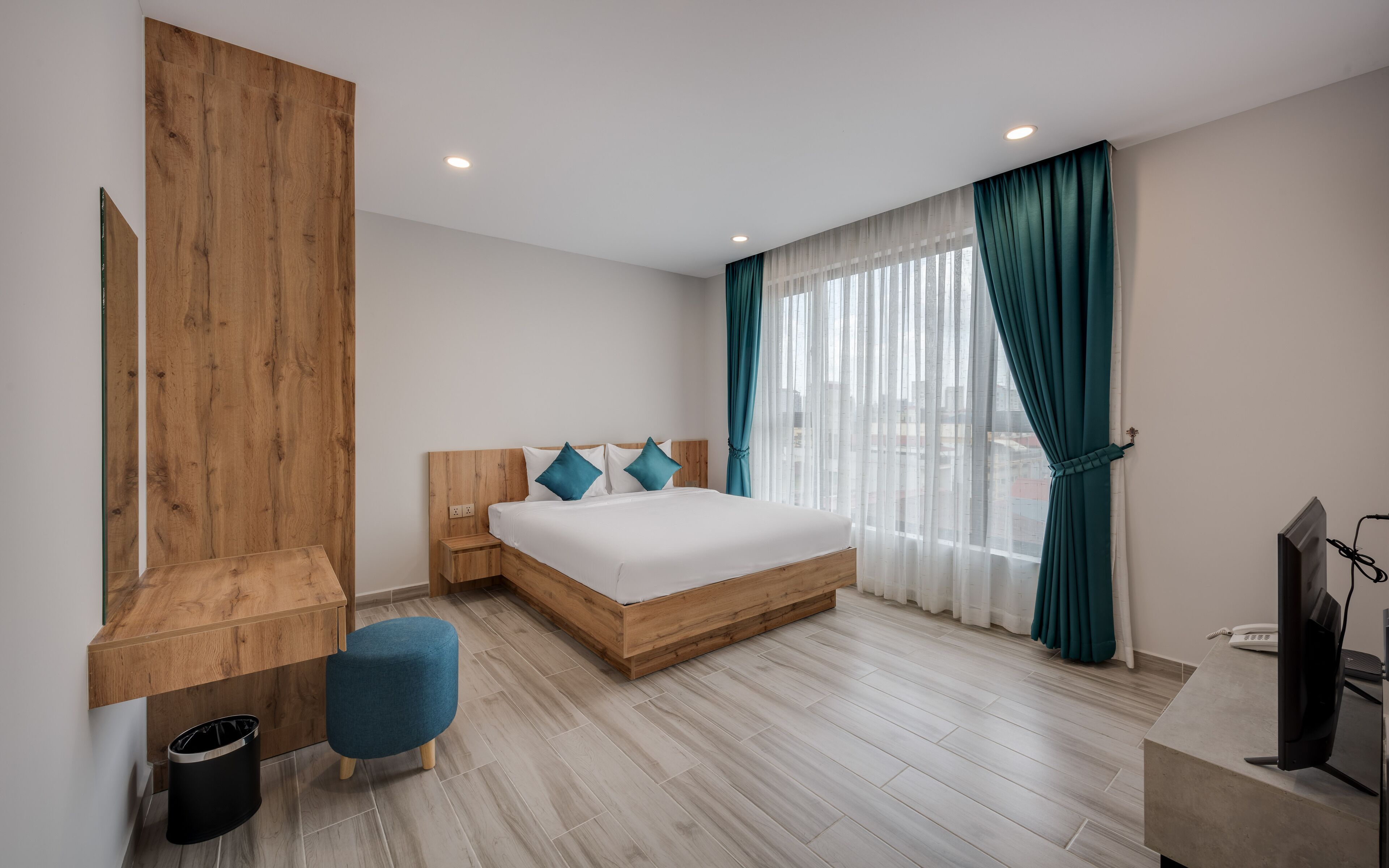 A modern bedroom with a wooden headboard, a large bed with white bedding, teal curtains, and a wooden nightstand with a trash bin.