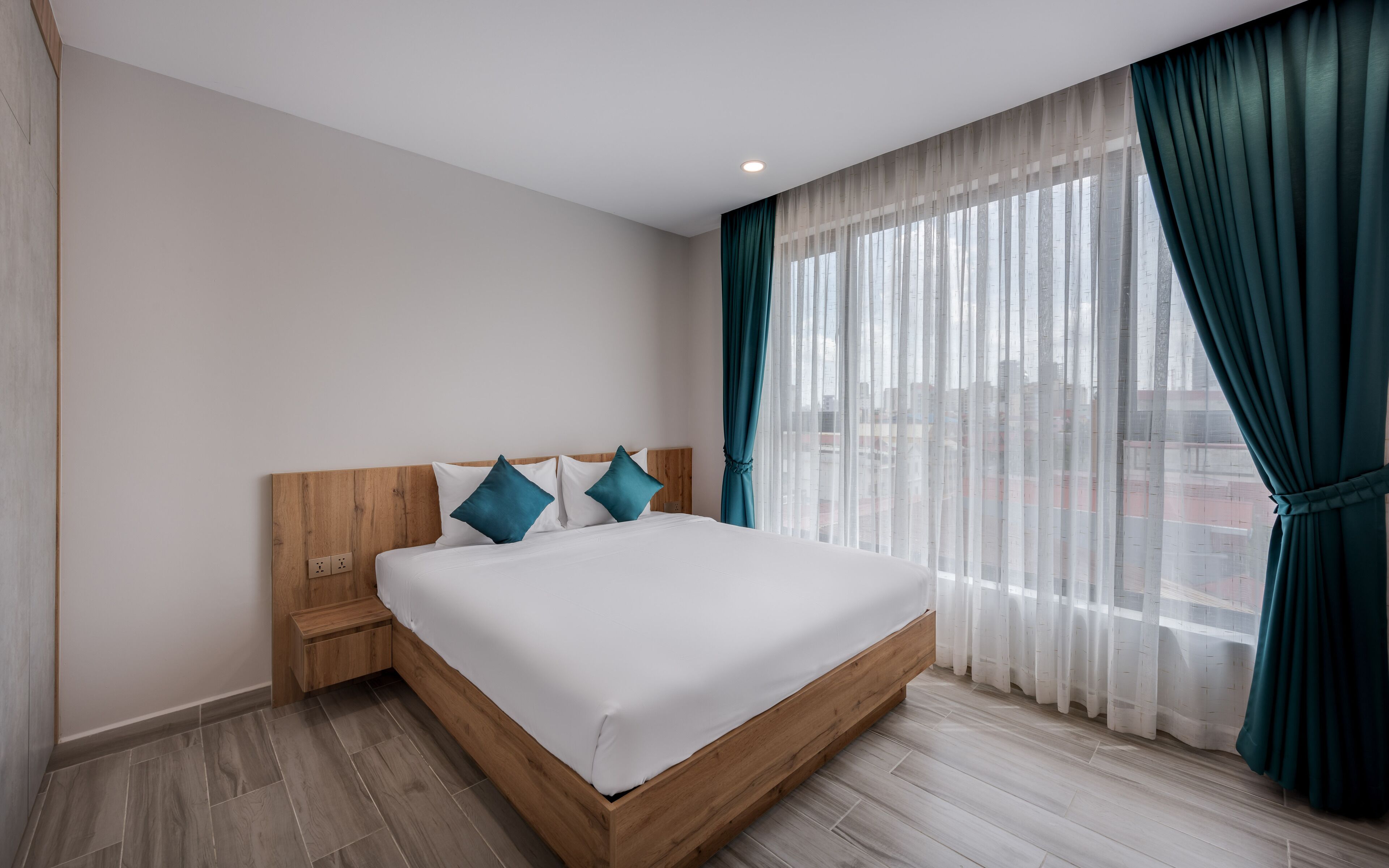 A modern hotel room with a wooden bed frame, white bedding, teal pillows, and sheer curtains.