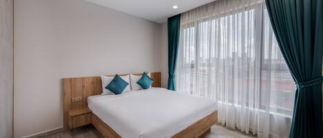 A modern hotel room with a wooden bed frame, white bedding, teal pillows, and sheer curtains.