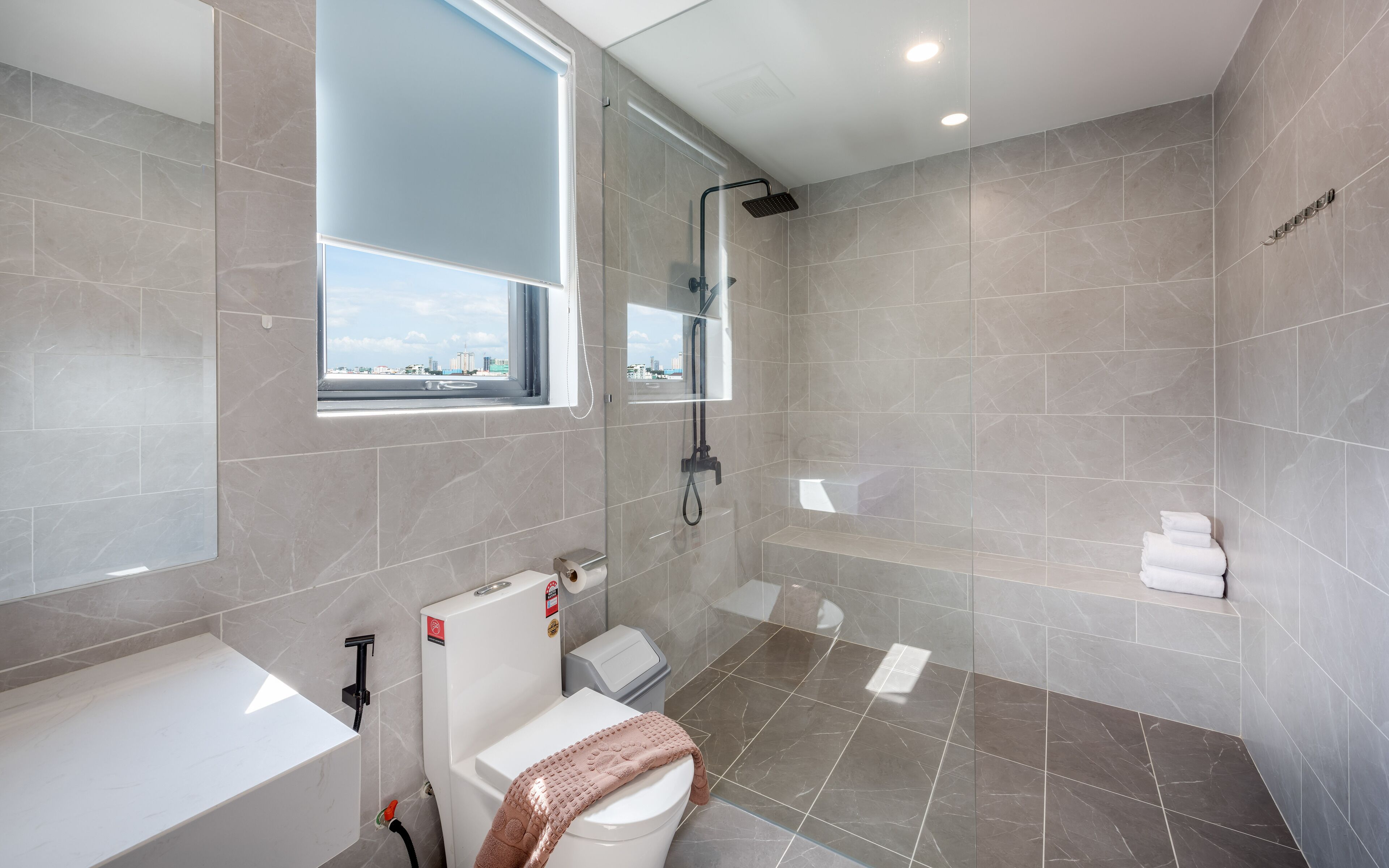 A modern bathroom with a shower, toilet, and sink, featuring grey tiles and a window with a city view.