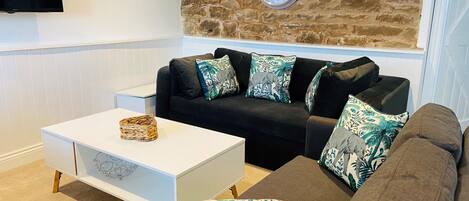 A living room with a white coffee table, a black sofa with patterned cushions, a stone wall, and a wall-mounted clock.
